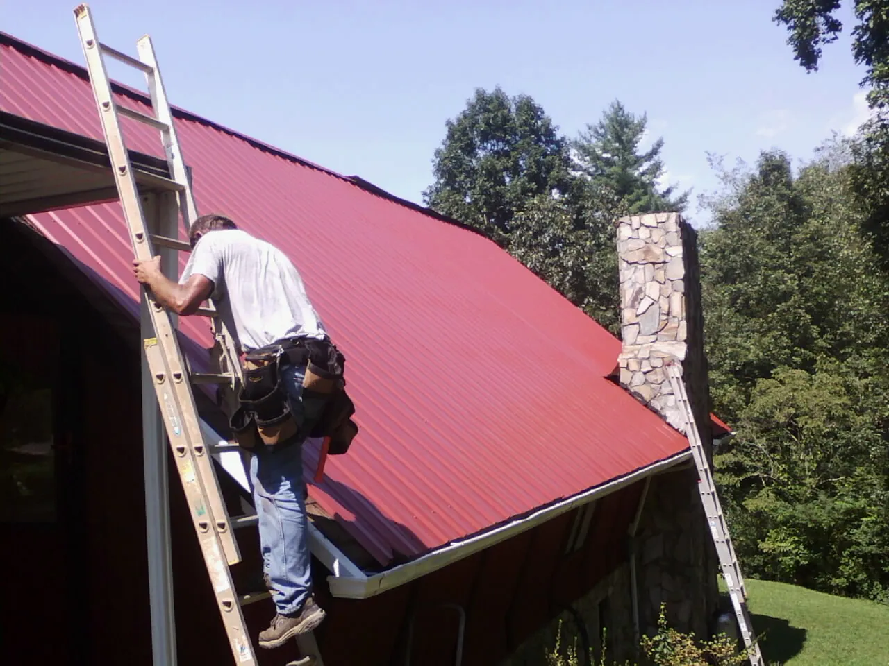 Roofer on a ladder beside a red metal roof and stone chimney — Soffit & Fascia Repair by Roofing Brothers in Chicora